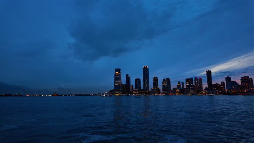 Blue hour skyline across Hudson River, Jersey City, New Jersey. Panoramic view of waterfront towers beneath heavy clouds with open water in the foreground.
