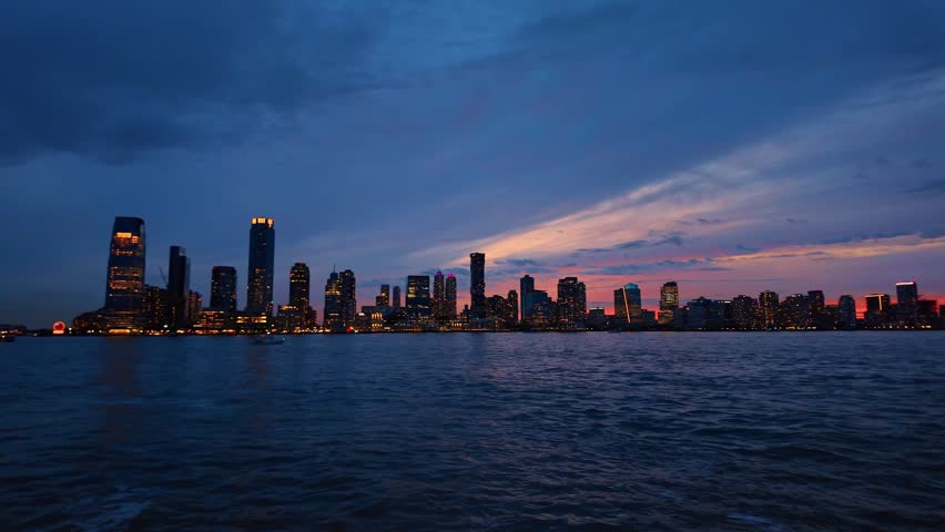 Wide skyline view across Hudson River, Jersey City, New Jersey. Distant waterfront towers and lights sit beneath layered blue clouds above rippled water.