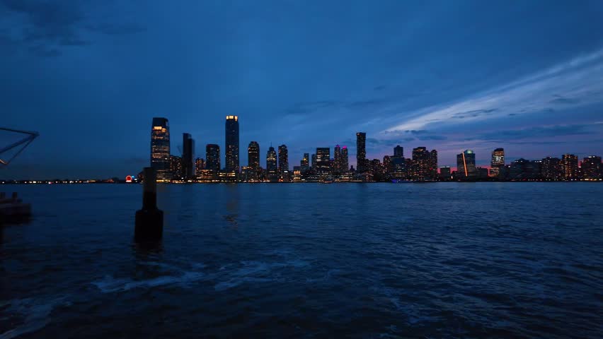 Ferry terminal facing Jersey City skyline, Hudson River, New Jersey. Illuminated canopy and pier posts form a foreground over dark water at blue hour.