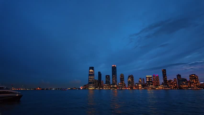 Waterfront view of Jersey City skyline, New Jersey. A passenger ferry crosses the Hudson River, with building lights reflected on evening water.
