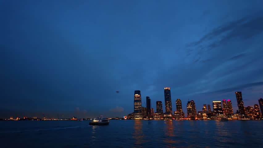 Distant passenger ferry below Jersey City towers, New Jersey. Wide view across the Hudson River with office lights, reflections and layered clouds.