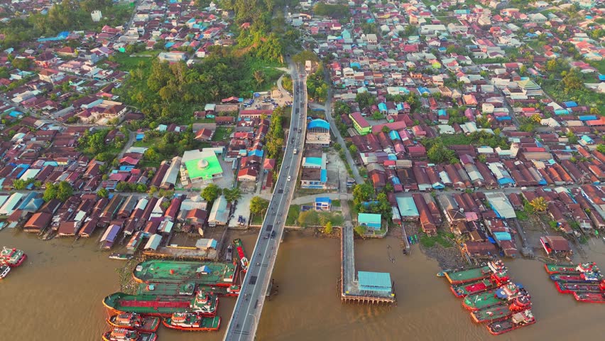 Aerial view of Mahkota Bridge spanning the river with surrounding residential rooftops and cargo ships docked along the muddy banks in Kecamatan Palaran, Kalimantan Timur, Indonesia.