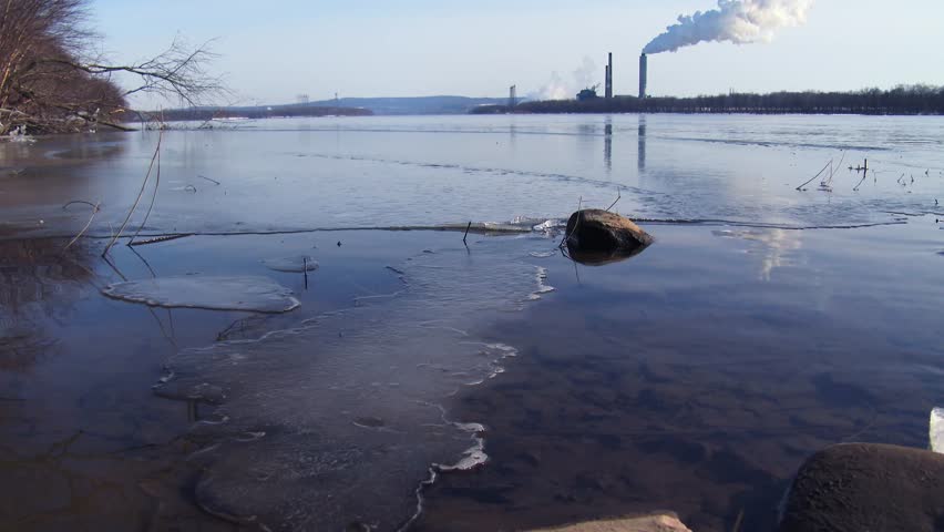 Footage of industrial area on a river bank with smoke and steam coming from factory pipes. The river is partially frozen, with remnants of ice along the shore, creating a stark image of environmental contrast.