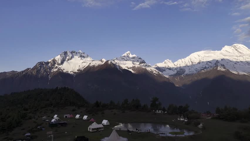 An breathtaking aerial view captures the vastness of the snow-capped Himalayan mountains in Tibet, China. Below, a serene campsite with tents and a small pond offers a tranquil scene against the