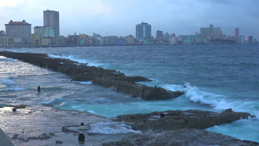 Dynamic ocean waves breaking over rocky coast line during cloudy day. Panoramic view of Havana cityscape across choppy water. Cinematic footage showing motion of sea and urban buildings.