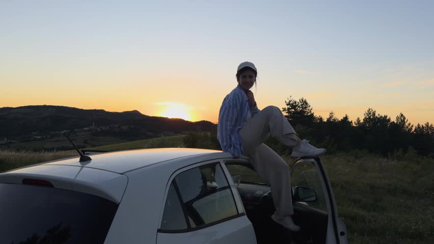 Young woman sitting on her car roof while admiring the beautiful golden hour sunset during a road trip in the italian countryside