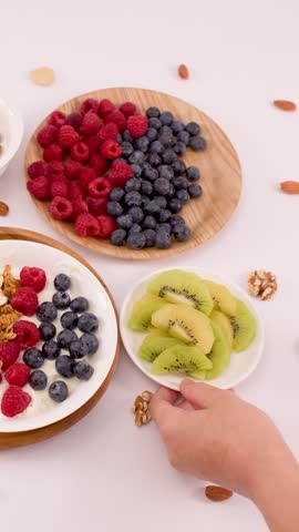 Hand adds sliced kiwi to a yogurt bowl with raspberries, blueberries, and crunchy granola.
