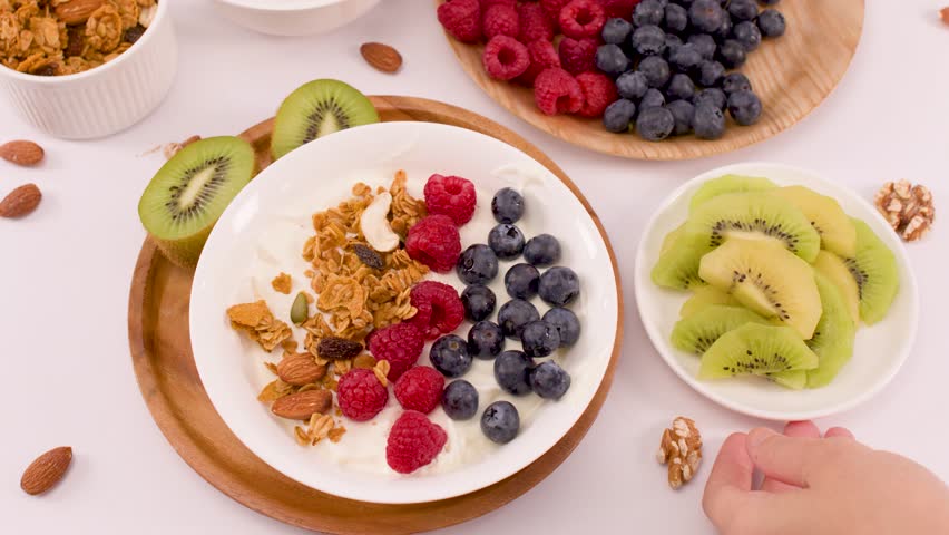 Overhead shot of hand placing kiwi slices beside yogurt bowl with berries and granola.