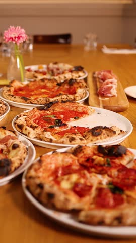 Hands reach for various gourmet pizzas on a restaurant table with wine and flowers.