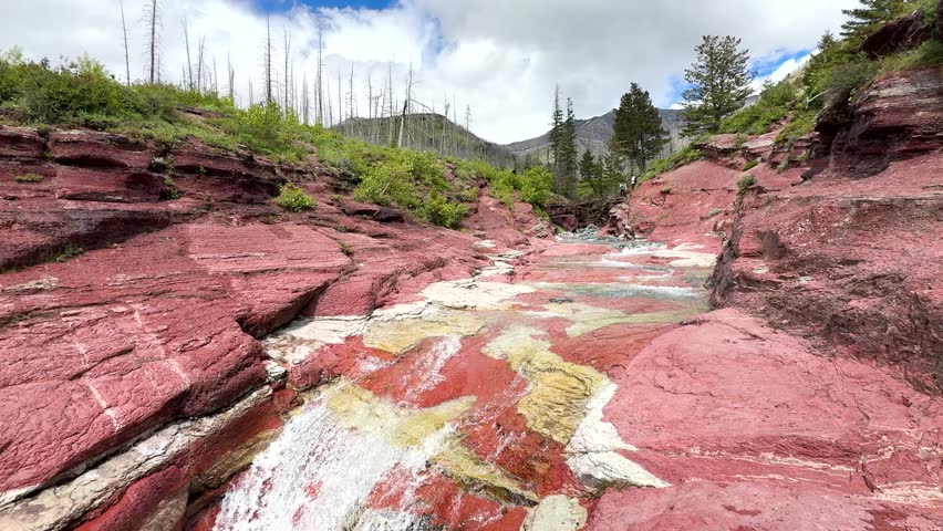 Red rock formations surrounded by green plants and trees. Water flows through the colorful rocks under a cloudy sky in a Tonwater national park in Canada.