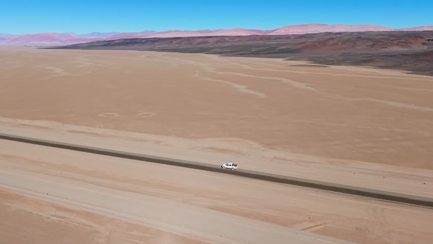 Aerial view of a lonely dirt road stretching through the arid desert landscape with a single vehicle driving along the path under a clear blue sky, Jujuy Province, Argentina.