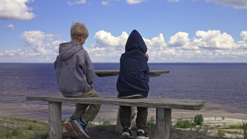 Two boys sit on a rustic wooden bench atop a grassy cliff, gazing across the vast blue river under a sky of fluffy clouds.