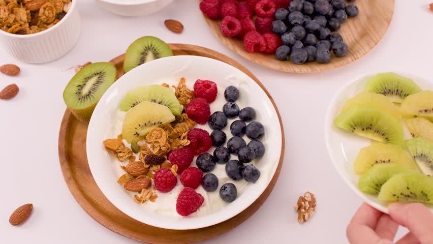 Overhead view of hands adding sliced kiwi to a yogurt bowl with berries and granola.