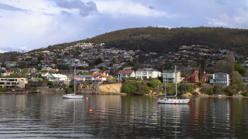 Coastal living and waterfront lifestyle are showcased with a housing development on a hillside overlooking Sandy Bay in Hobart, Australia. Sailboats are anchored in the calm water, reflecting the sky