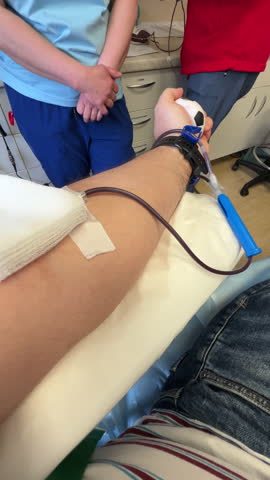 A close up point of view shot showing a patient donating blood while squeezing a stress ball, with nurses in scrubs supervising the procedure.