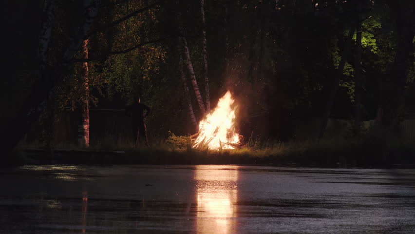 Sparkling bonfire on lake shore at campsite at night, man looking at mesmerizing flame of campfire
