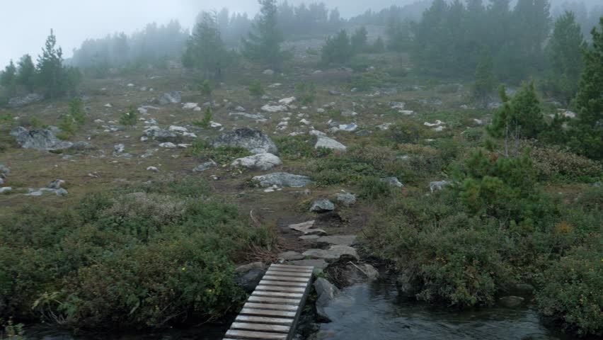 A static handheld shot of a small wooden footbridge crossing a tranquil mountain stream. The rocky landscape is covered in green shrubs and pine trees, shrouded in heavy morning fog and mist.