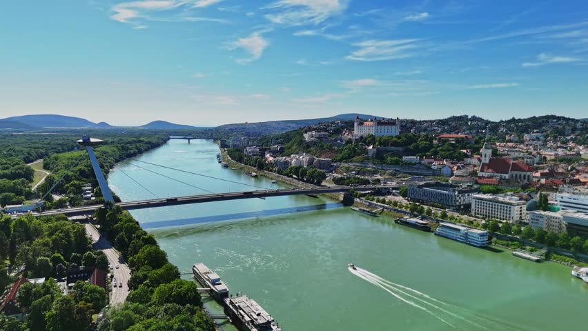 People enjoy the daytime in Bratislava, Slovakia. The Danube River flows through the city with boats moving. Buildings line the riverbank under a clear sky.