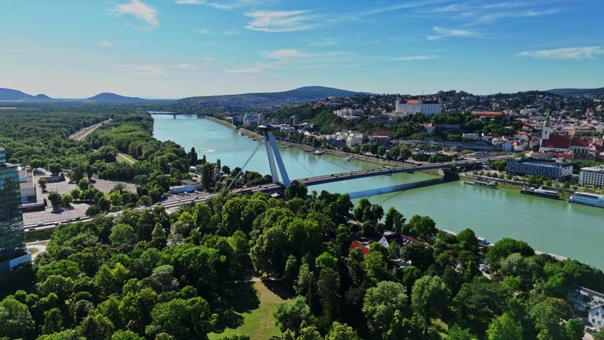 The Danube River flows through Bratislava, Slovakia, showing green parks and buildings along the shore. A bridge connects both sides of the river.