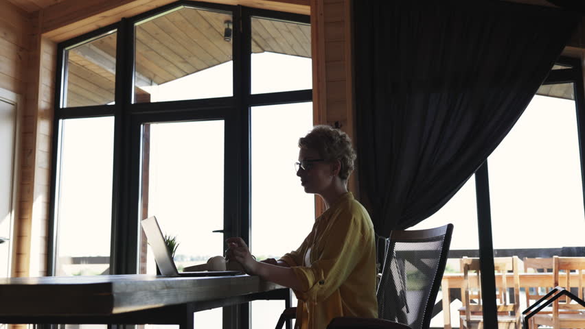 A woman with short curly hair and glasses waves hello while participating in a video conference call on her laptop in a wooden cabin