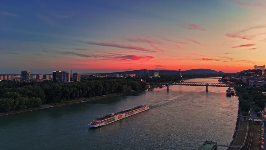 A boat travels along the Danube River during sunset in Bratislava. The sky shows warm colors as buildings line the riverbank.