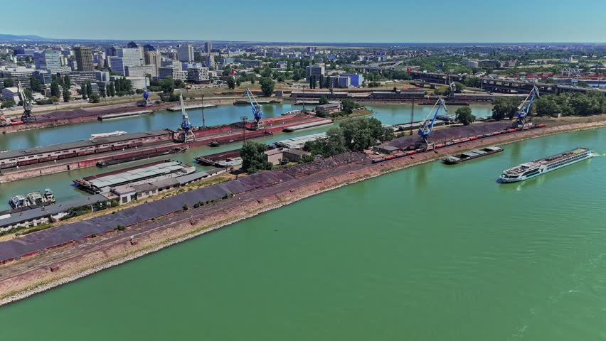 Ships and cranes are seen at the docks along the Danube river in Bratislava. The city skyline is visible in the background under clear blue skies.