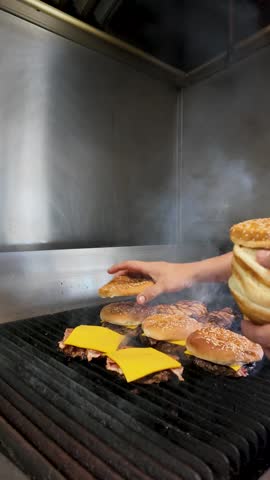Short order cook’s hands making fresh bacon cheeseburgers on a smoky commercial grill at a roadside diner. Melting cheese, sizzling bacon, and classic buns - close up vertical
