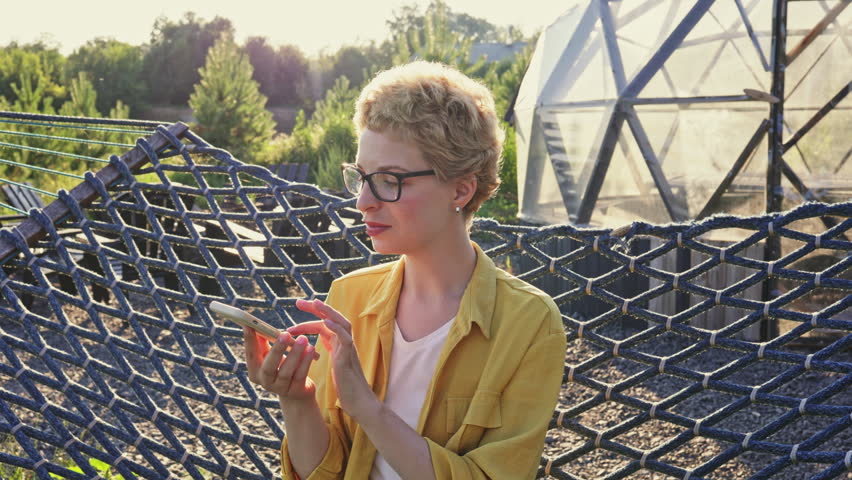 A young caucasian woman with short blonde hair and glasses is sitting on a hammock and talking on her smartphone