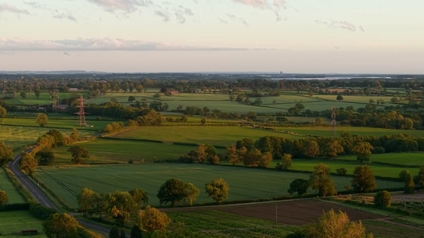 Green farmland landscape with country road and hedgerows. Aerial view of agricultural fields divided by trees and rural lanes. Peaceful countryside panorama stretching across rolling farmland at