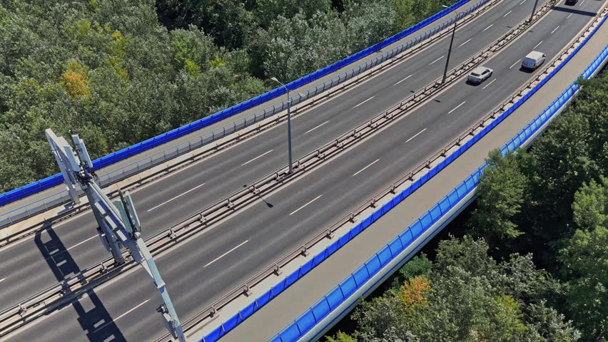 Cars drive along a road surrounded by trees in Bratislava, Slovakia. The road has a blue fence on one side. The setting shows typical urban infrastructure.