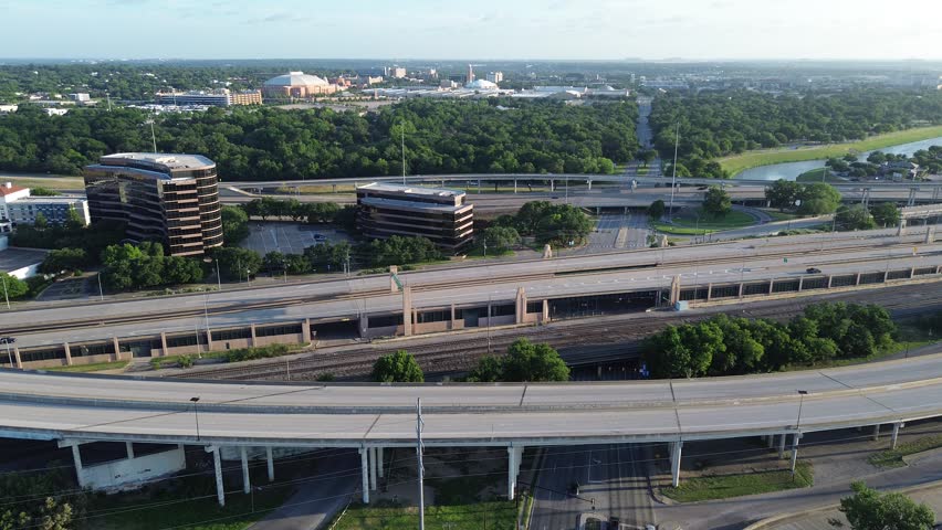 Elevated highways slice through horizon behind corporate office buildings, densely forested areas. Morning sun casts elongated shadows across linear railroad tracks below, Fort Worth, Texas. USA