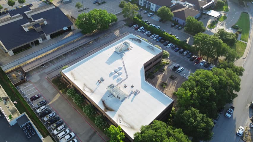 White roofs featuring HVAC units dominate commercial complex surrounded by dense tree canopies. Parking rows flank office building as morning light highlights urban footprint, Fort Worth, Texas. USA