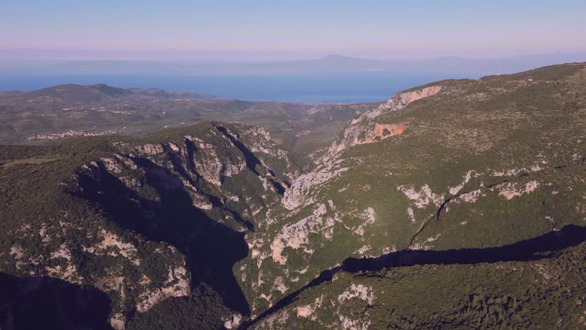 Aerial drone footage showing a deep mountain gorge in the Peloponnese, Greece. The landscape features steep limestone cliffs, rugged rocky slopes, dense Mediterranean vegetation, forested hills, deep shadows, and a dramatic ravine cutting through the mountains. The blue sea and distant coastline are visible on the horizon, adding a coastal atmosphere to the wild mountain scenery. Suitable for projects about Greek nature, rugged landscapes, mountain geography, outdoor exploration, hiking regions, wilderness, scenic travel, Mediterranean cliffs, and natural formations in southern Greece.