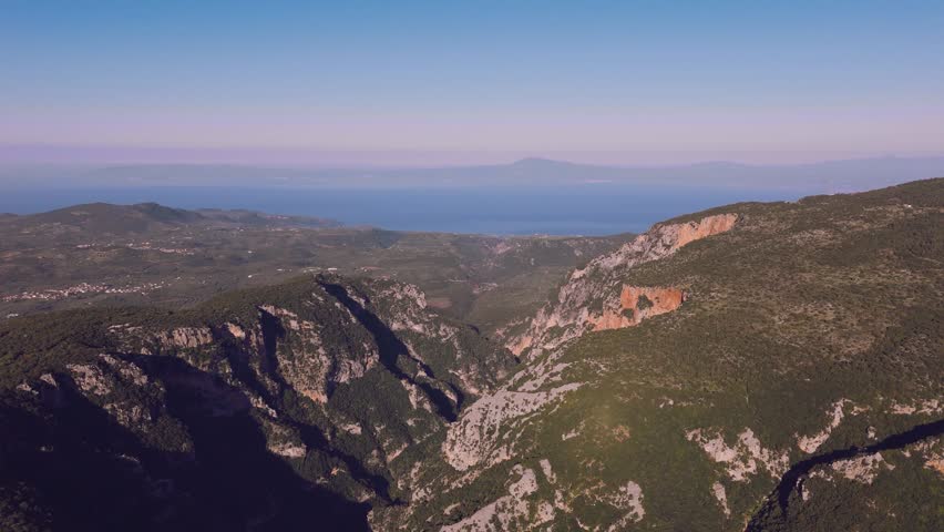 Aerial drone footage showing a deep mountain gorge in the Peloponnese, Greece. The landscape features steep limestone cliffs, rugged rocky slopes, dense Mediterranean vegetation, forested hills, deep shadows, and a dramatic ravine cutting through the mountains. The blue sea and distant coastline are visible on the horizon, adding a coastal atmosphere to the wild mountain scenery. Suitable for projects about Greek nature, rugged landscapes, mountain geography, outdoor exploration, hiking regions, wilderness, scenic travel, Mediterranean cliffs, and natural formations in southern Greece.