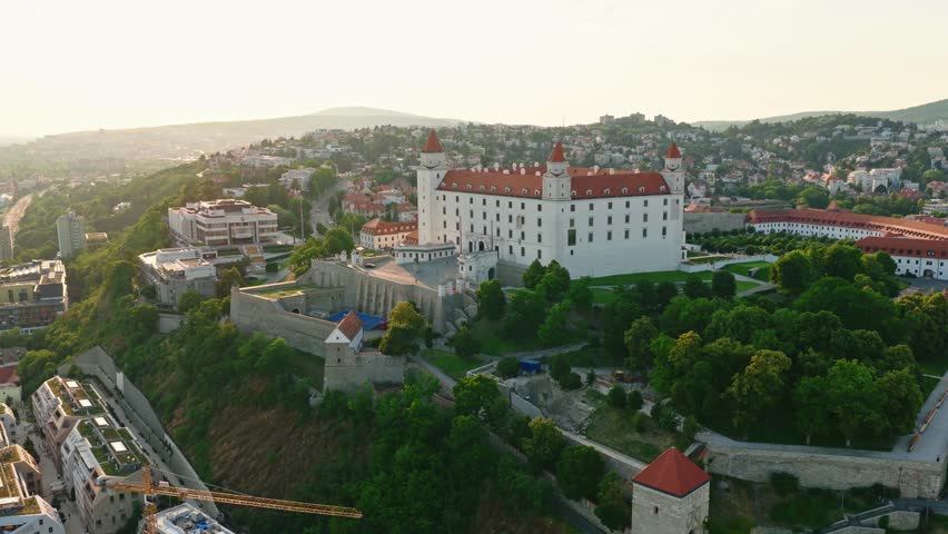 Drone aerial view of Bratislava Castle during golden hour sunset. Historic landmark and symbol of Bratislava, Slovakia, surrounded by greenery and urban cityscape with scenic evening light.
