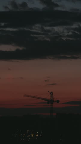 Timelapse of a tall construction crane silhouette against a dramatic, colorful sky during sunset, with dark clouds moving over a city with glowing lights in the background at twilight