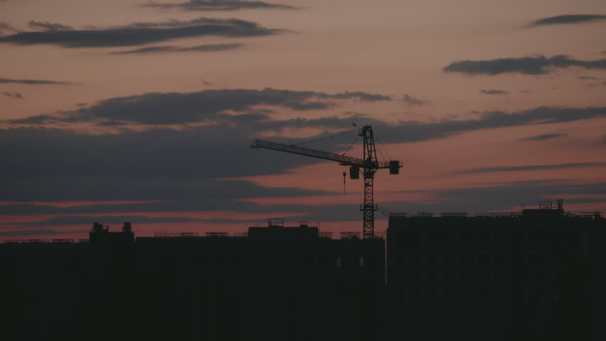 Dramatic timelapse showing a construction crane silhouette working against a beautiful evening sky with colorful clouds moving over the dark cityscape at dusk, highlighting urban development