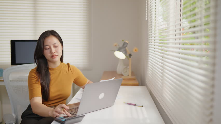Woman working on a laptop at a home office while reviewing documents, ideal for remote work, online business, administration, planning, productivity and workflow concepts.