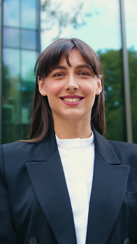 Successful young businesswoman standing outdoors, smiling and looking at the camera confidently, with glass office in the background. Slow motion