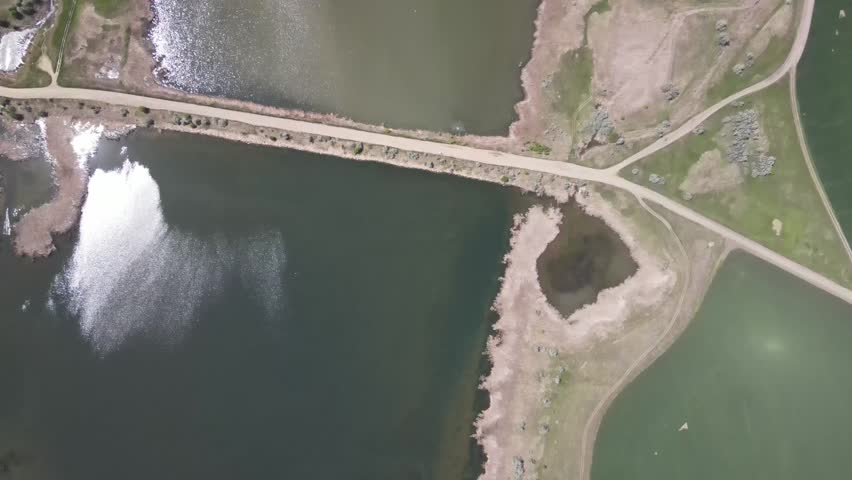 Aerial drone footage showing a top-down view of a cascade of ponds connected by a dam embankment. The footage captures the water system