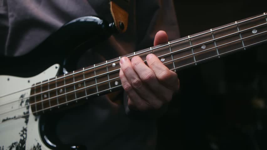 Close up skilled bass player hand fretting notes on neck of black and white electric bass guitar, studio recording session in dark environment. Musician playing vintage bass guitar fretboard