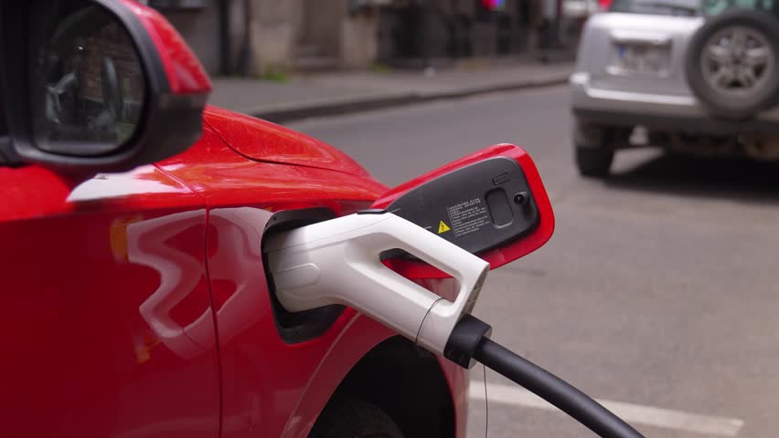 Close-up view of a modern red electric vehicle connected to a charging station, refueling its battery on a bustling city street with traffic passing by in the background on a daytime