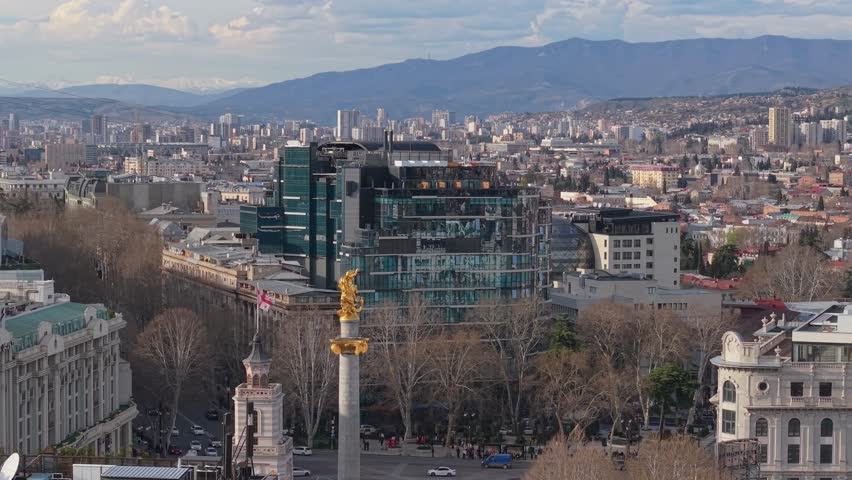 Orbiting shot of the golden St George statue at Freedom Square, revealing the surrounding Tbilisi cityscape with dynamic urban perspective.