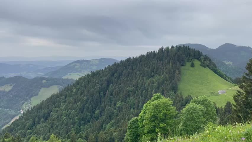 Panoramic view of a scenic mountain landscape with lush green meadows, steep rocky cliffs, and a large lake surrounded by distant mountains under a cloudy sky.