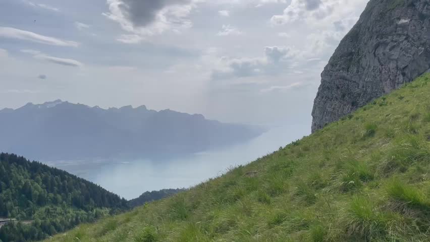 Panoramic view of a scenic mountain landscape with lush green meadows, steep rocky cliffs, and a large lake surrounded by distant mountains under a cloudy sky.