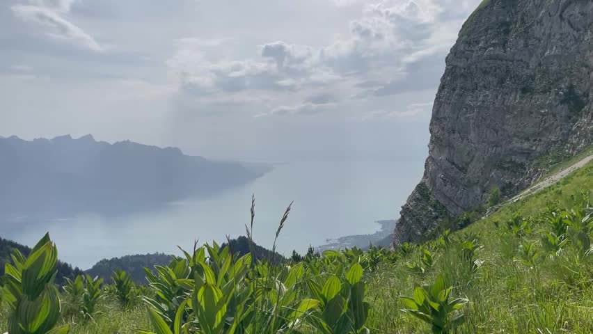 Panoramic view of a scenic mountain landscape with lush green meadows, steep rocky cliffs, and a large lake surrounded by distant mountains under a cloudy sky.