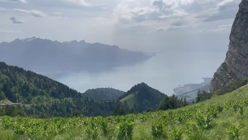 Panoramic view of a scenic mountain landscape with lush green meadows, steep rocky cliffs, and a large lake surrounded by distant mountains under a cloudy sky.