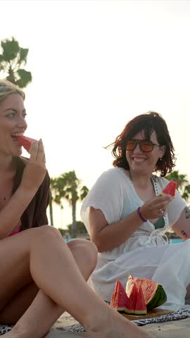 Two smiling female friends enjoying a fresh watermelon picnic on the beach during a beautiful sunset, laughing together and sharing a happy moment on their summer vacation getaway by the sea