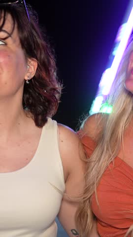 Two beautiful female friends enjoying a fun night out at an amusement park, smiling and laughing together with a brightly lit ferris wheel illuminating the dark background behind them