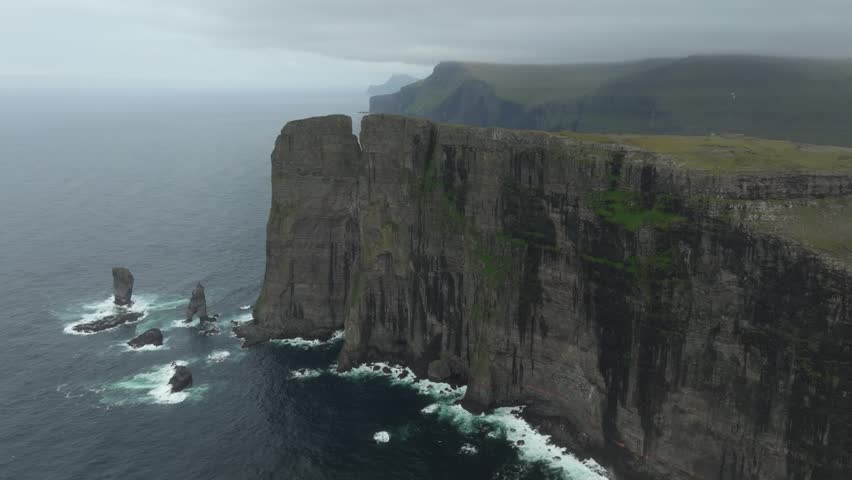 Aerial drone shot panning left over the majestic cliffs of Eiðiskollur mountain, near Eiði, on the Faroe Islands. Featuring rugged coastlines, a vast ocean, and misty, cloudy skies.